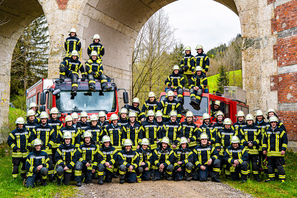 Die Mannschaft der Feuerwehr Stiefenhofen posiert in Einsatzmontur mit Helm vor zwei Feuerwehrautos, einige Personen sitzen auf den Autos. Im Hintergrund sind die Bögen einer Brücke zu sehen.