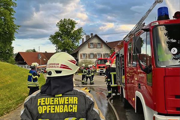 Zu sehen ist ein Feuerwehrmann von hinten mit der Aufschrift "Feuerwehr Opfenbach" auf seiner Jacke. Ebenso ist ein Feuerwehrauto rechts angeschnitten und es sind noch weitere Feuerwehrmänner von weitem sichtbar.